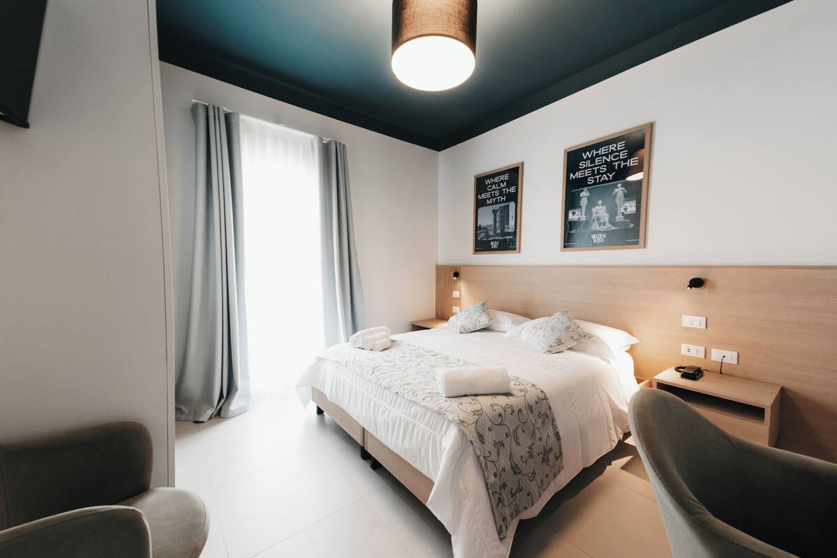 Una habitación de hotel moderna con una cama grande, una mesita de noche, una silla y una ventana con cortinas.