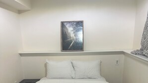 Ein ordentlich bezogenes Bett in einem Zimmer mit einem einzelnen Wandbild, einem kleinen Tisch und einem gemusterten Vorhang.