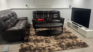 Ein Wohnzimmer mit einer Ledersofa, einem Fernsehsessel, einem Couchtisch, einem Fernseher und einem flauschigen Teppich.