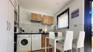 A compact kitchen with a washing machine, wooden cabinets, a glass-top dining table, and white chairs. - Seafoam - T1 BIS near the ocean (Mimizan)