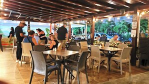 Un ristorante all'aperto con tavoli e sedie, una zona coperta e vista sulla strada con auto e palme.