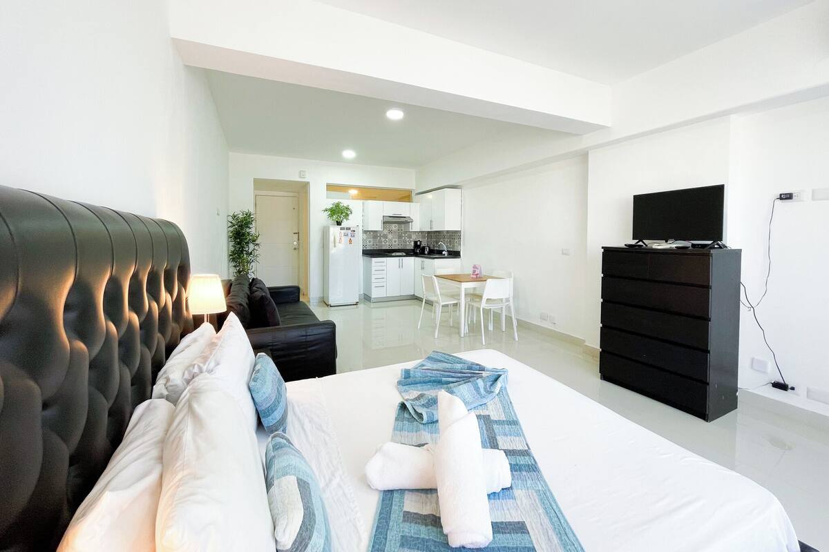 Une chambre d’hôtel moderne avec un grand lit, une télévision, une commode et, au fond, une petite cuisine.