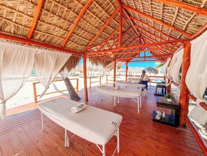 Een houten terras met rieten dak, witte bedden en een massagetafel.