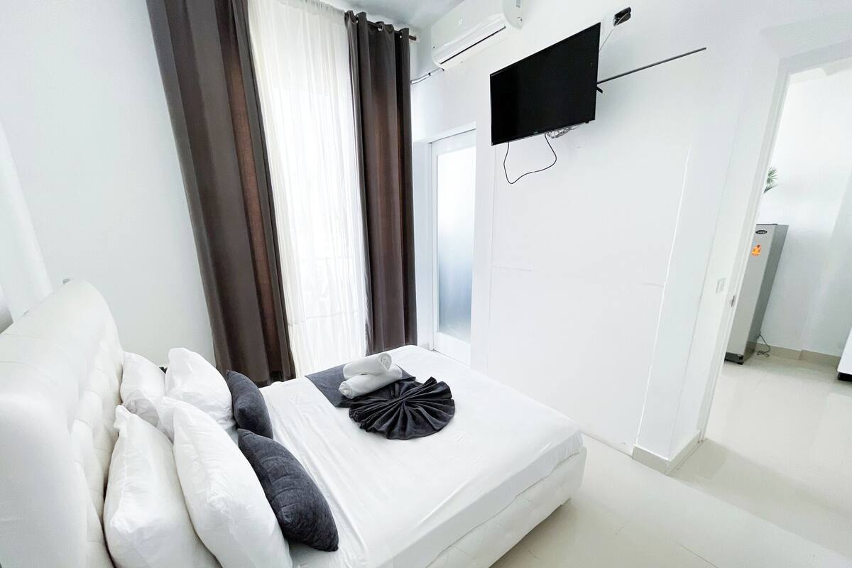 Une chambre d’hôtel moderne avec un lit, des murs blancs, un téléviseur fixé au mur et une fenêtre avec des rideaux.