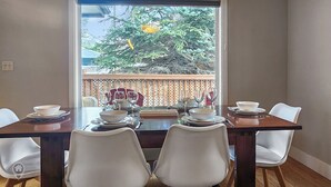 A dining table set for four with white chairs, a view of a tree through a window, and a wooden deck outside.