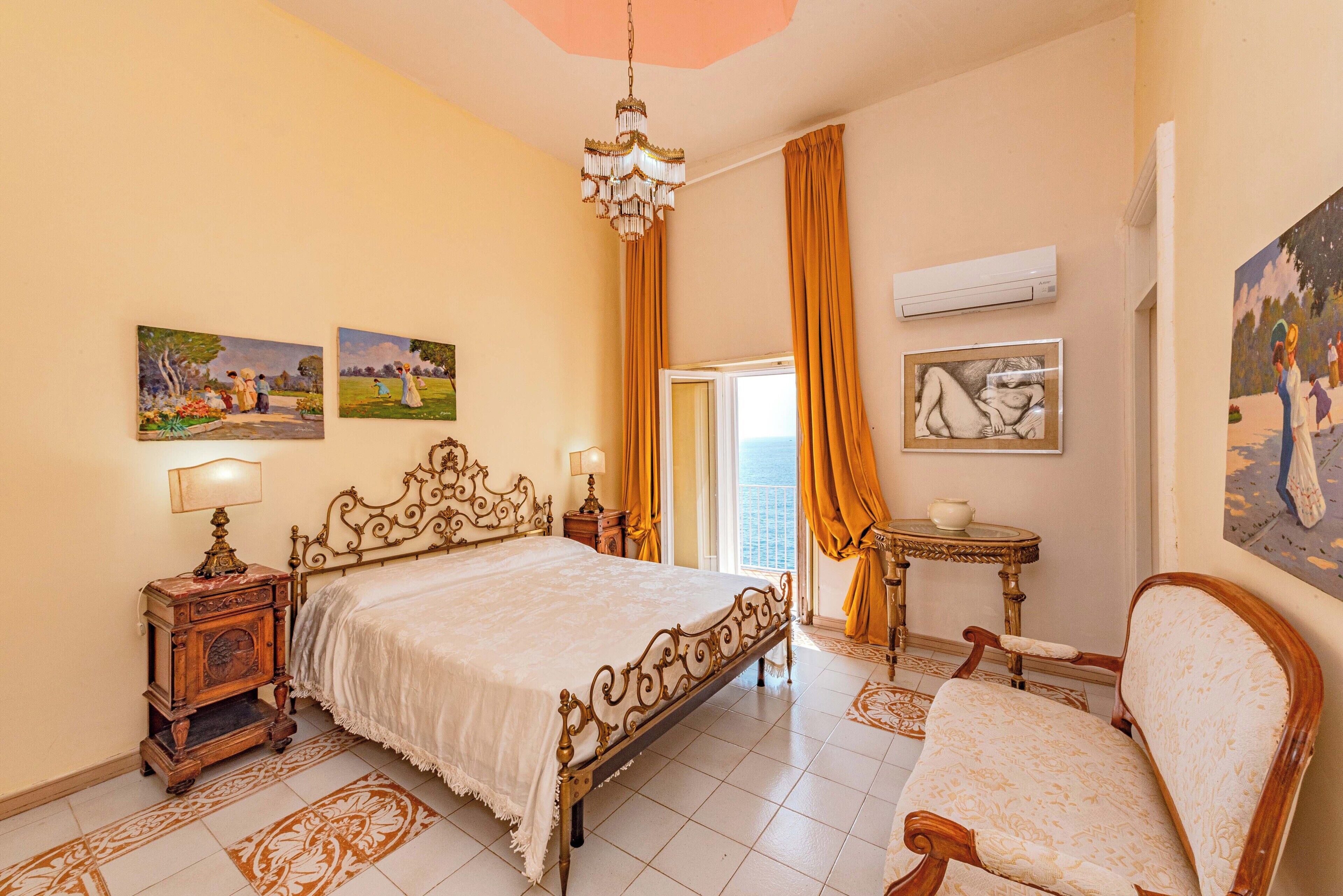 A bedroom with a bed, a chair, a nightstand, a chandelier, and artwork on the walls.