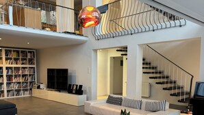 Un amplio salón con una mesa de centro de vidrio, un sofá blanco, una estantería y una escalera.