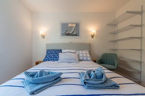 Ein ordentlich bezogenes Bett mit blau-weißer Bettwäsche, zwei Wandlampen, ein gerahmtes Bild über dem Bett und ein grüner Sessel neben einem Metallregal.