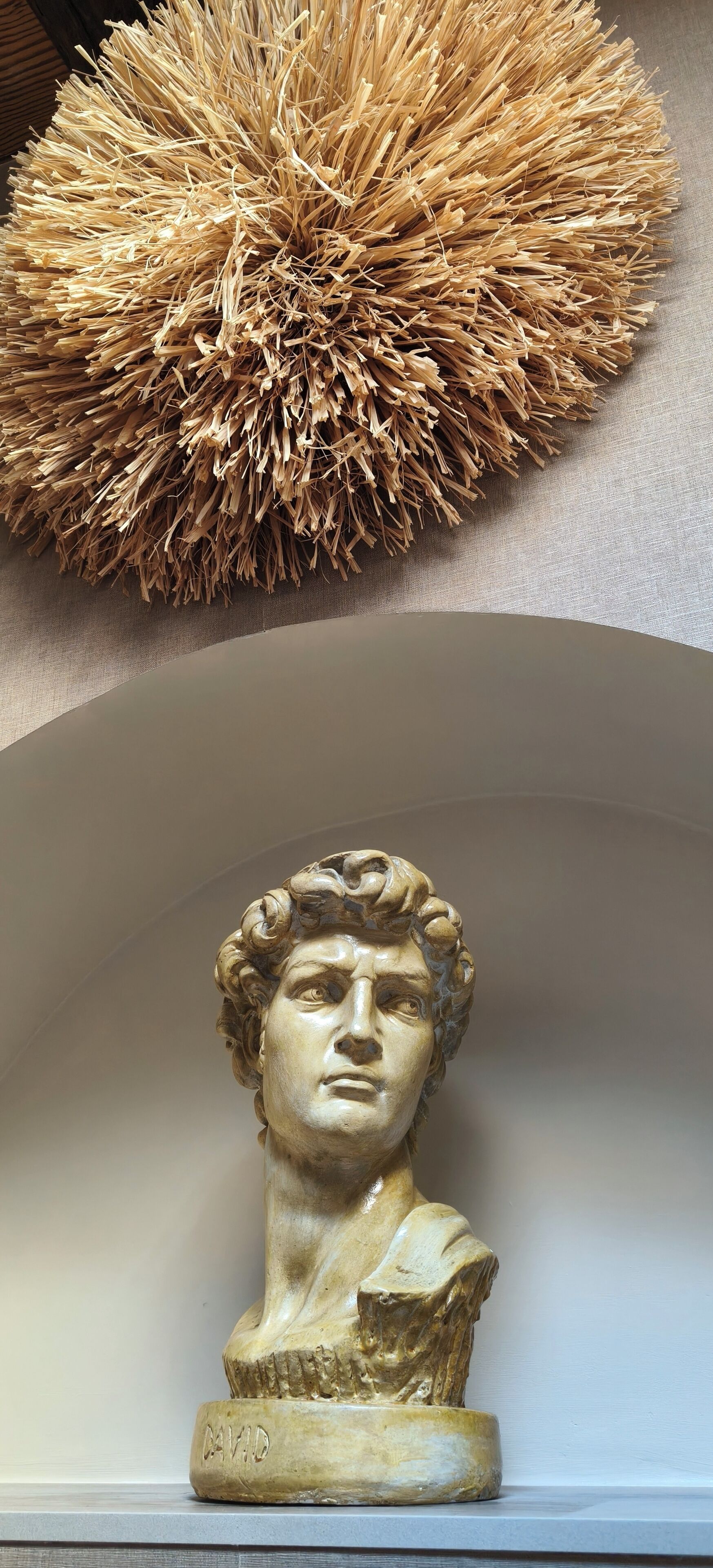 En skulptur föreställande en byst, placerad på en piedestal som är märkt med texten ”DAVID”, i ett rum med en vägg som har en tydlig struktur och ett runt, strukturerat föremål ovanför.