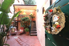Um corredor exterior estreito com escadas, plantas em vasos e uma grinalda decorativa numa porta.