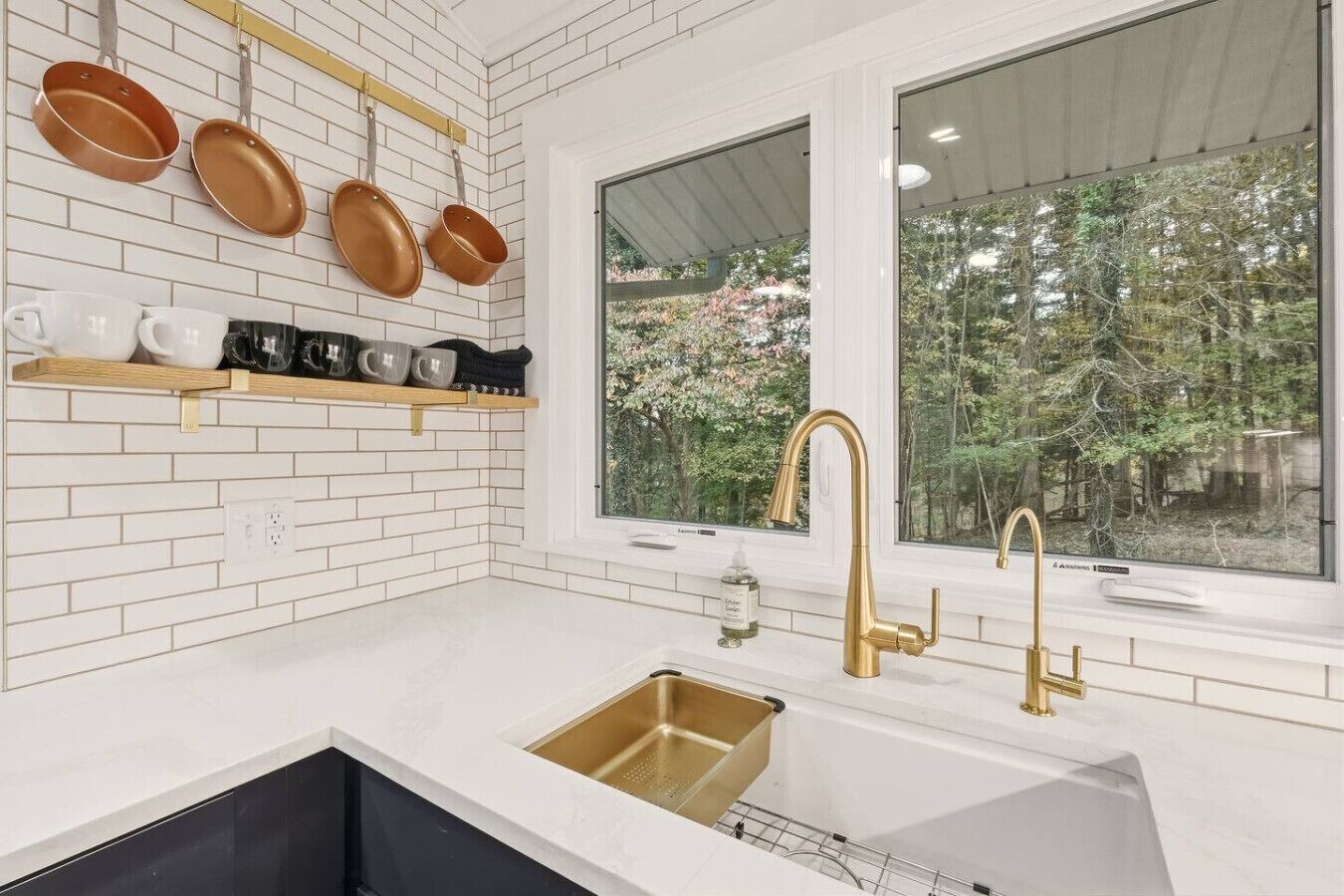 Una cocina con azulejos blancos tipo metro, un grifo dorado y una ventana con vistas a árboles.