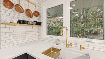 Una cocina con azulejos blancos tipo metro, un grifo dorado y una ventana con vistas a árboles.