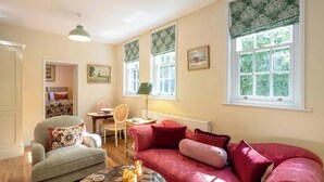 Et hyggeligt rum med sofa, stol, bord og lampe. Der er vinduer med gardiner og en radiator.