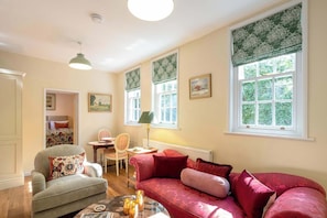 A cozy room with a sofa, a chair, a table, and a lamp. There are windows with curtains and a radiator. - Osbaston Mews - Stableyard Cottage (Market Bosworth)