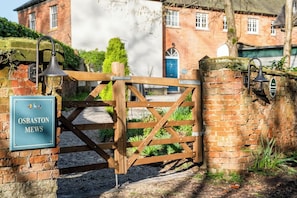 A wooden gate with a sign reading 'OSBASTON MEWS' and a brick wall with a lamp post. - Osbaston Mews - Stableyard Cottage (Market Bosworth)