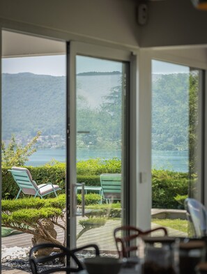 Blick durch eine Glastür auf eine Terrasse mit Gartenmöbeln, einer Steinskulptur und einen See mit Bergen im Hintergrund.