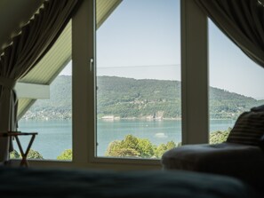 Ein Zimmer mit großem Fenster und Blick auf einen See und einen bewaldeten Hügel.