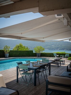 Ein Poolbereich mit Tisch und Stühlen, eine überdachte Terrasse und Blick auf Wasser und Hügel.