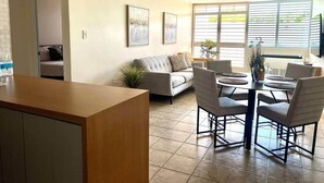 Een woonkamer met een bank, een eettafel met stoelen, een keukenhoek en een groot raam met jaloezieën.