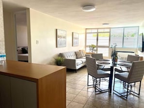 Een woonkamer met een bank, een eettafel met stoelen, een keukenhoek en een groot raam met jaloezieën.