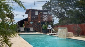 Una piscina con muelle de madera, aro de baloncesto y un edificio con balcón al fondo.