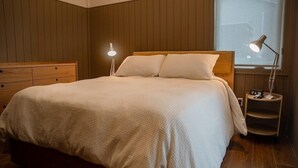 A bedroom with a wooden headboard, a bed with a white quilt, two bedside lamps, and a nightstand with a phone and a lamp. - Superior Queen Two-Bedroom Cottage (Bronson)