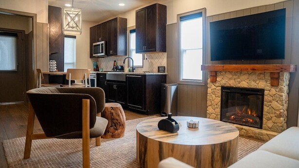 A modern living room with a fireplace, a kitchen with dark wood cabinets, a flat-screen TV, and a round coffee table. - Superior Queen Two-Bedroom Cottage (Bronson)