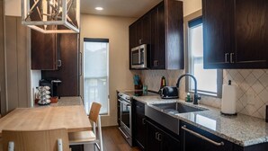 A modern kitchen with dark wood cabinets, a stainless steel microwave, a double sink, and a wooden dining table. - Superior Queen Two-Bedroom Cottage (Bronson)