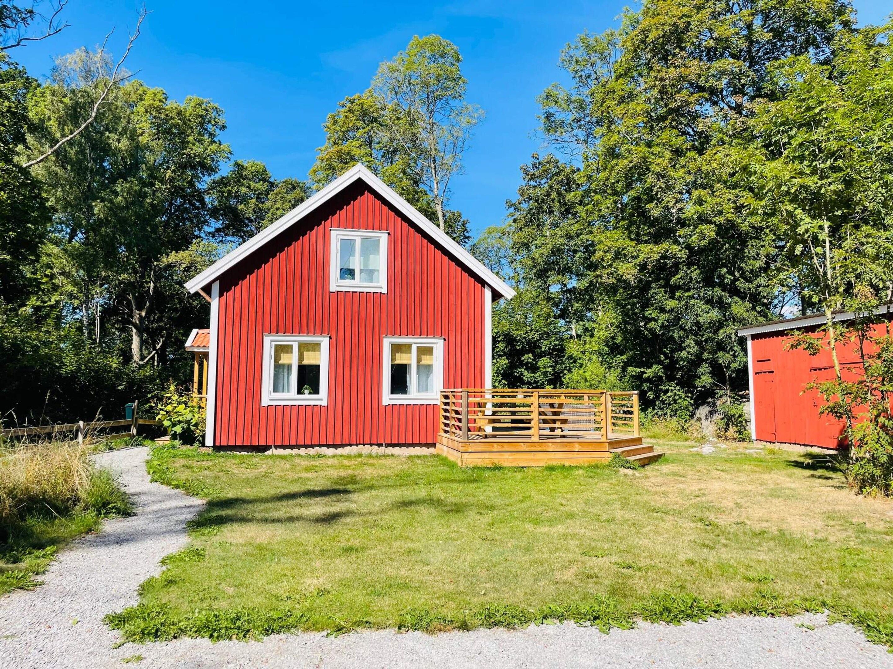 A red wooden cabin with a gravel path leading to it, surrounded by greenery.