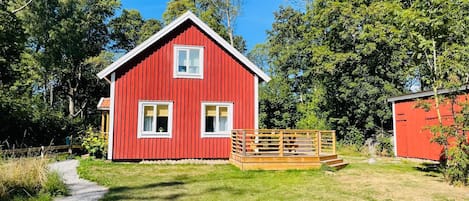 A red wooden cabin with a gravel path leading to it, surrounded by greenery.
