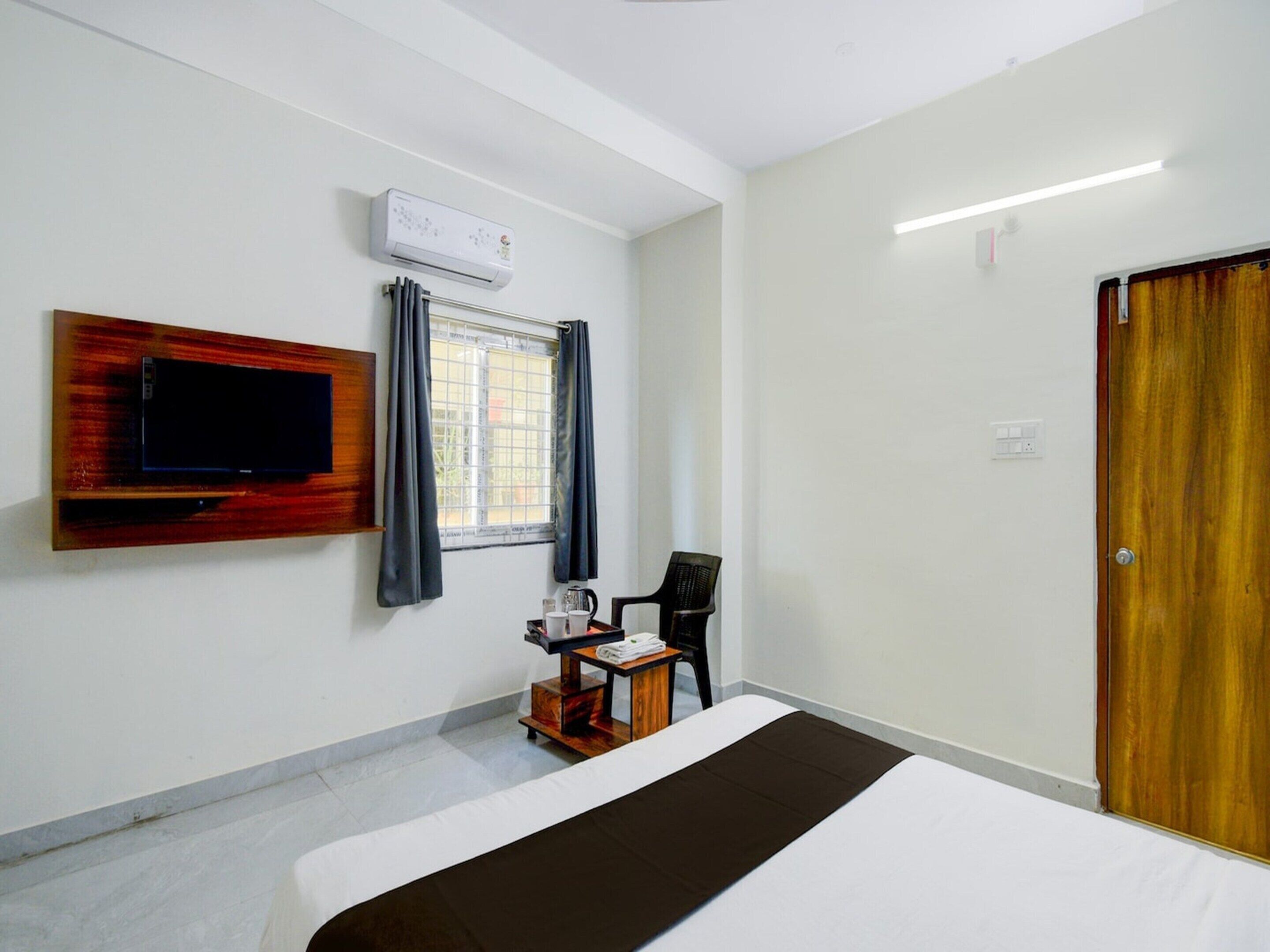 A hotel room with a bed, a TV mounted on the wall, a window with curtains, a chair, and a wooden door.