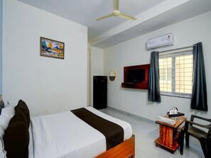 Camera d'hotel con un letto, una TV a muro, una ventola a soffitto, un condizionatore d'aria, una finestra con tende e un tavolino con una sedia.