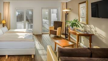 Habitación de hotel con cama, sofá, sillón, mesa, televisor y balcón con barandilla.