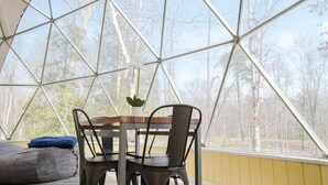 A geodesic dome structure with a table and chairs inside, a potted plant on the table, and a view of trees through the transparent ceiling.