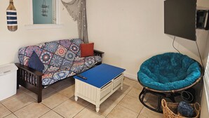 A room with a patterned sofa, a blue ottoman, a teal chair, a ceiling fan, and a TV mounted on the wall.