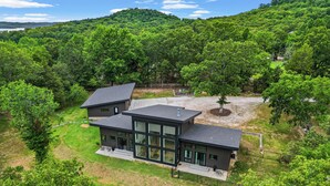 A modern two-story house with large windows, surrounded by lush greenery and a hilly landscape.