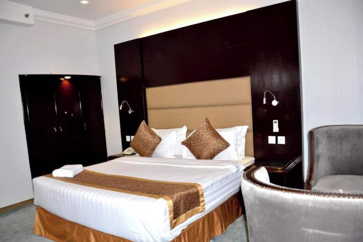 A hotel room with a large bed, a chair, a wardrobe, and a bedside table.