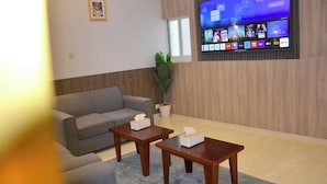 A living room with a grey sofa, two wooden tables with tissues, a potted plant, and a mounted TV displaying various channels. - Ferbion Hotel Abha (Abha)