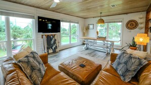 Ein geräumiges Wohnzimmer mit Holzbalkendecke, Ledersofas, einem Fernseher und großen Fenstern mit Blick auf Grünflächen.