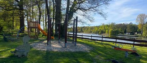 Un parque infantil con columpios y tobogán, rodeado de árboles y vistas a un lago.