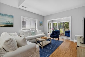 Salón con sofá blanco, una mesita de centro y una silla azul. Hay ventanas con persianas, un cuadro en la pared y vistas al exterior.