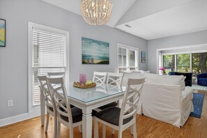 A dining area with a glass table and white chairs, a living area with a sofa and a blue chair, and a chandelier. - Award-Winning Resort Amenities | Links Clubhouse 10 (Isle Of Palms)