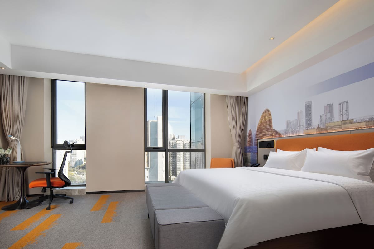 Una habitación de hotel con una cama grande, un escritorio, una silla de oficina, una ventana con vista a la ciudad y un mural urbano en la pared.