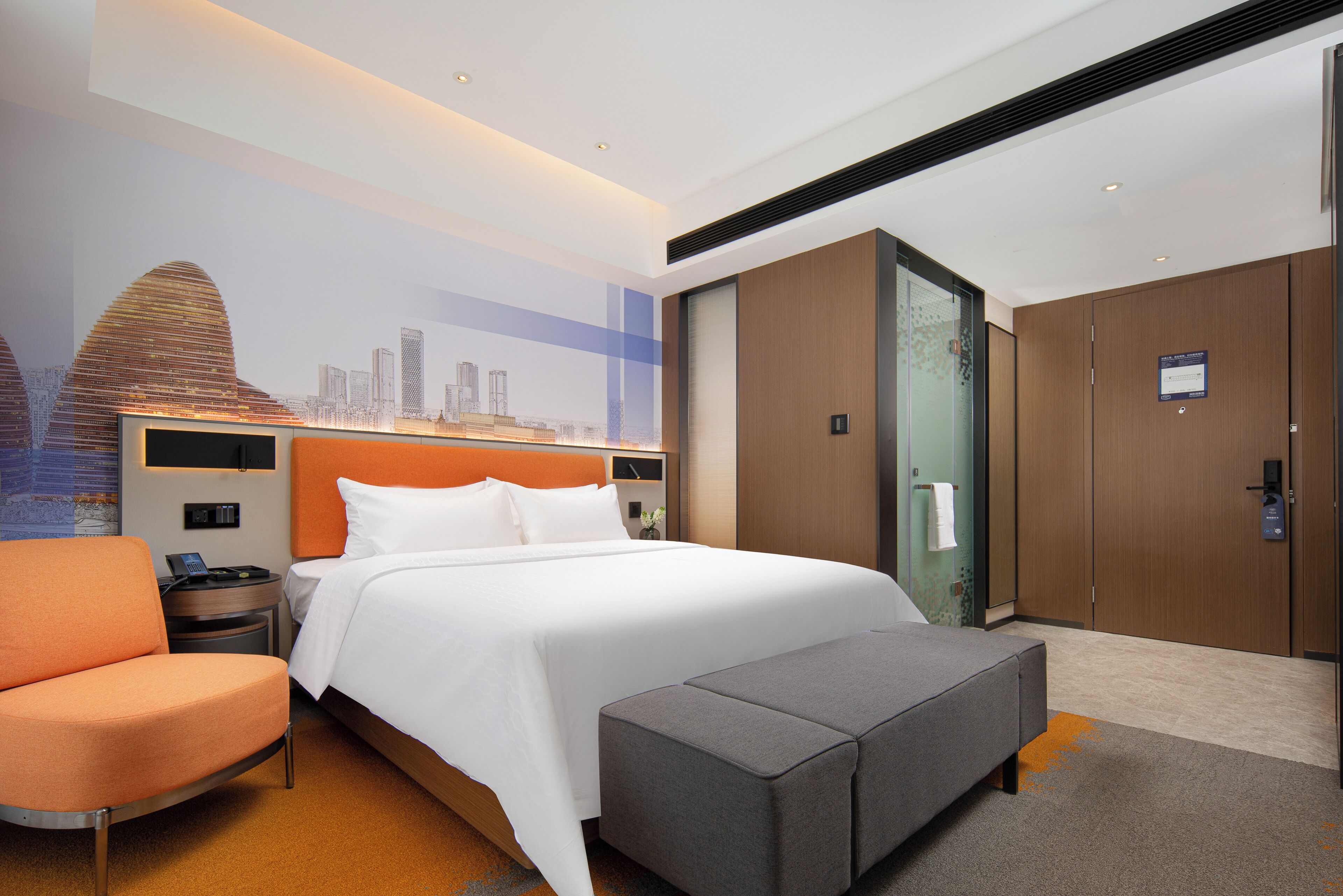 Una habitación de hotel con cama, una silla naranja, un reposapiés gris, una mesita de noche y un mural con vista a la ciudad.