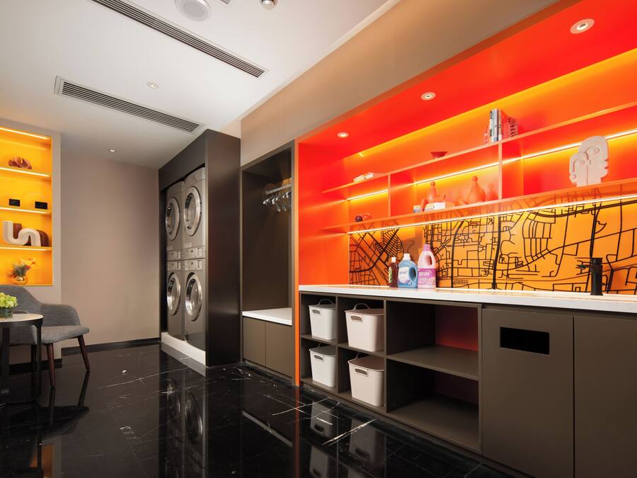 Un baño moderno con piso de mármol negro, un sofá gris, una lavadora y un tocador naranja muy iluminado con repisas.