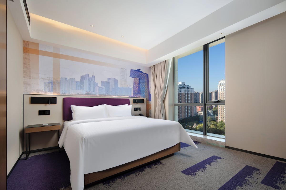 Una habitación de hotel con una cama grande, vista a la ciudad, un mural del horizonte urbano y una ventana con cortinas.