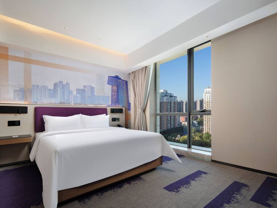Una habitación de hotel con una cama grande, vista a la ciudad, un mural del horizonte urbano y una ventana con cortinas.