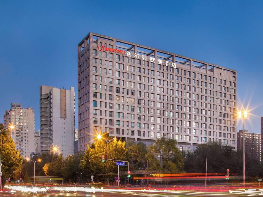 Un hotel de varios pisos con fachada de diseño cuadriculado, múltiples ventanas y un cartel que indica que es un hotel Hampton.