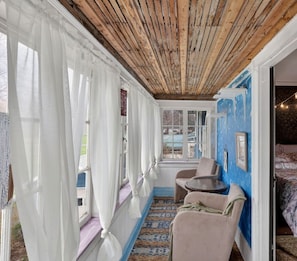 Ein Zimmer mit Holzboden, einer Fensterbank mit Vorhängen, einem runden Tisch und einem Bett an einer blauen Wand.