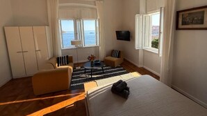 A hotel room with a bed, a sofa, a TV, a window with a view of the sea, and a chandelier. - Onyx Apartments 2&4 (Dubrovnik)